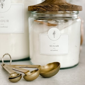 May include: A glass jar with a wooden lid filled with white sugar. The jar has a label that says "Sugar - all natural". There are four gold measuring spoons in the foreground.