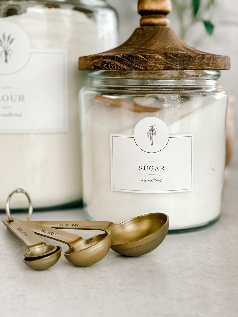 May include: A glass jar with a wooden lid filled with white sugar. The jar has a label that says "Sugar - all natural". There are four gold measuring spoons in the foreground.