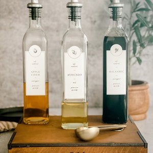 Puede incluir: Tres botellas de vidrio con boquillas para verter y etiquetas para vinagre de sidra de manzana, aceite de aguacate y vinagre bals&aacute;mico. Las botellas est&aacute;n sobre una caja de madera con la palabra "ACEITE" grabada en la parte delantera. Dos cucharas medidoras de metal dorado est&aacute;n delante de las botellas.