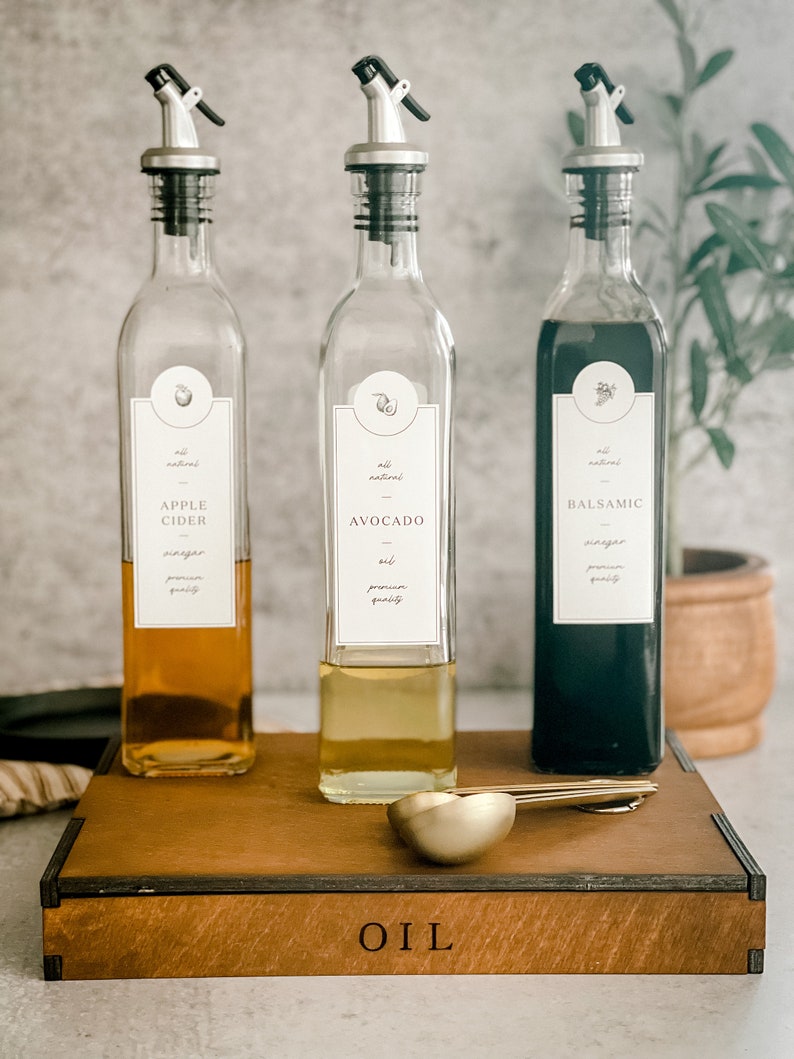Puede incluir: Tres botellas de vidrio con boquillas para verter y etiquetas para vinagre de sidra de manzana, aceite de aguacate y vinagre bals&aacute;mico. Las botellas est&aacute;n sobre una caja de madera con la palabra "ACEITE" grabada en la parte delantera. Dos cucharas medidoras de metal dorado est&aacute;n delante de las botellas.