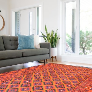 May include: A gray sofa with a blue pillow sits in front of a large window with a view of trees. A potted plant is next to the sofa. A colorful patterned rug covers the floor.