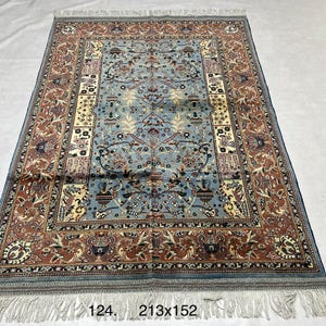 Puede incluir: Una alfombra rectangular que presenta un campo central azul claro con intrincados motivos florales y de urnas, rodeado por un borde ancho de color terracota con patrones detallados. La alfombra incluye flecos blancos en ambos extremos y mide 213x152 cm.