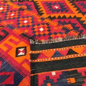 May include: A traditional Turkish rug with a geometric pattern in shades of red, orange, black, and blue. The rug has a fringe border and is woven with intricate details.