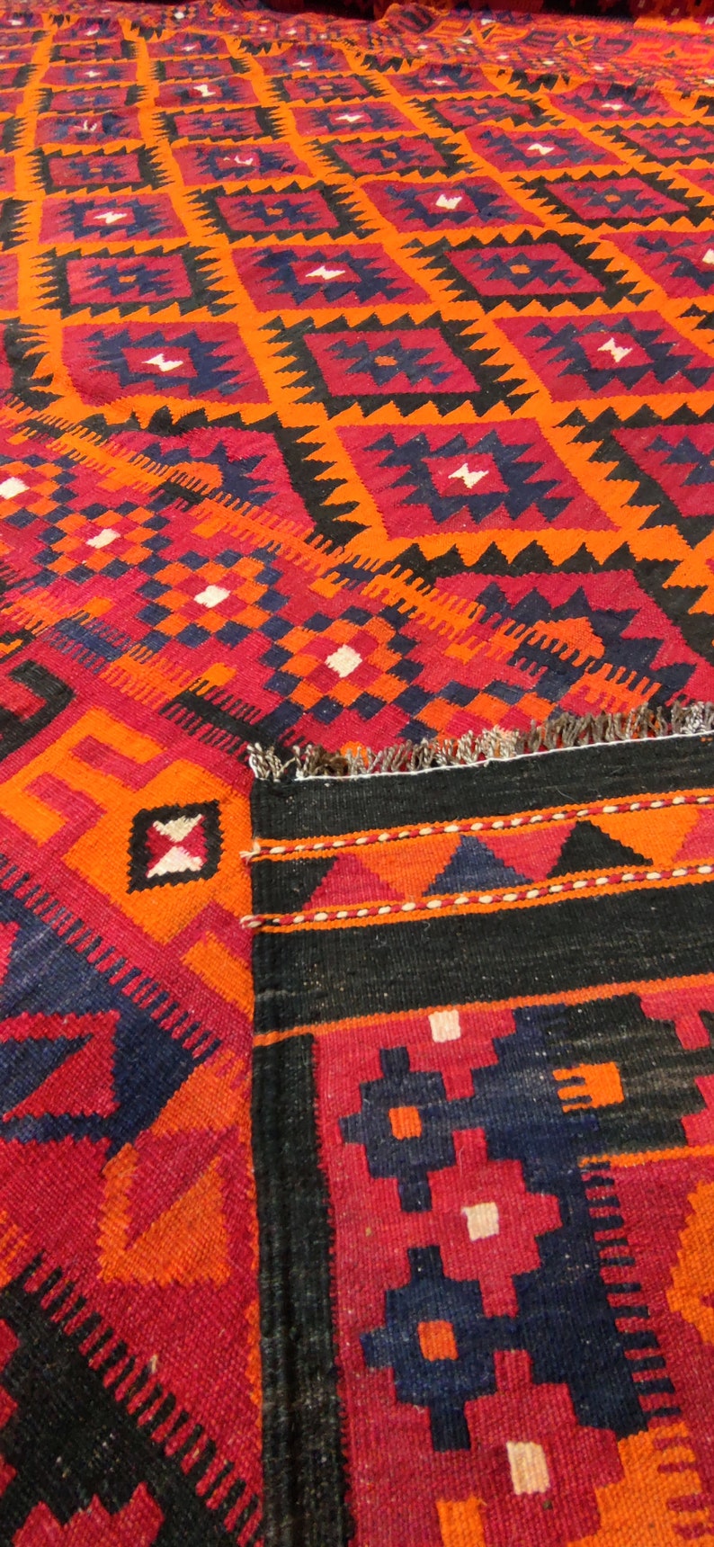 May include: A traditional Turkish rug with a geometric pattern in shades of red, orange, black, and blue. The rug has a fringe border and is woven with intricate details.
