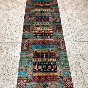 K&ouml;nnte beinhalten: Ein langer, schmaler Teppich mit einem farbenfrohen, geometrischen Muster. Der Teppich ist aus Wolle und hat an beiden Enden Fransen. Die Farben des Teppichs sind Rot, Blau, Gr&uuml;n, Gelb und Braun.