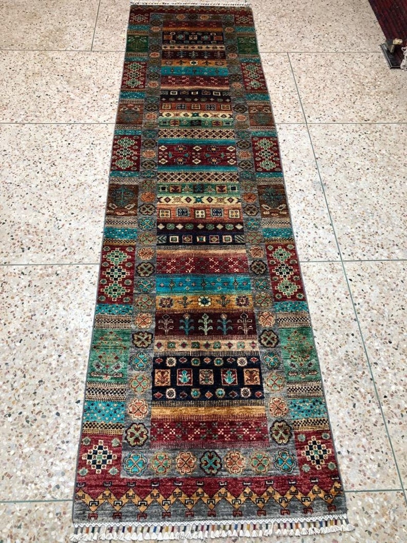 K&ouml;nnte beinhalten: Ein langer, schmaler Teppich mit einem farbenfrohen, geometrischen Muster. Der Teppich ist aus Wolle und hat an beiden Enden Fransen. Die Farben des Teppichs sind Rot, Blau, Gr&uuml;n, Gelb und Braun.