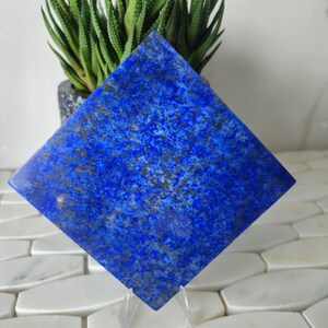 Könnte beinhalten: Ein quadratisches Stück polierter Lapislazuli mit einer tiefblauen Farbe und goldenen Flecken. Der Stein wird auf einem klaren Acryl-Ständer präsentiert.