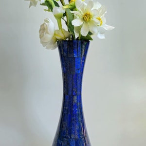Könnte beinhalten: Eine hohe, blaue Mosaikvase mit weißen Blumen und gelben Zentren und grünen Stielen. Die Vase hat einen ausgestellten oberen und unteren Teil mit einem schmaleren Mittelteil. Das Mosaik besteht aus kleinen, rechteckigen blauen Steinen.