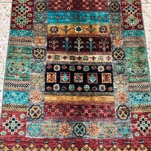 K&ouml;nnte beinhalten: Ein bunter Teppich mit einem geometrischen Muster in Rot-, Blau-, Gr&uuml;n- und Goldt&ouml;nen. Der Teppich hat einen Fransenrand und ist aus Wolle gefertigt.