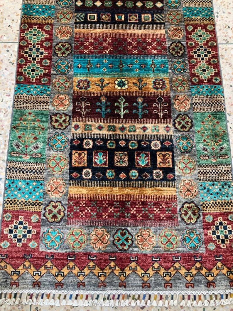 K&ouml;nnte beinhalten: Ein bunter Teppich mit einem geometrischen Muster in Rot-, Blau-, Gr&uuml;n- und Goldt&ouml;nen. Der Teppich hat einen Fransenrand und ist aus Wolle gefertigt.