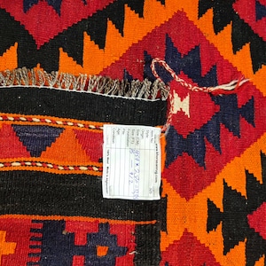 May include: A close-up of a colorful rug with a geometric pattern in shades of orange, red, black, and blue. The rug has a fringe border and a price tag attached to it.