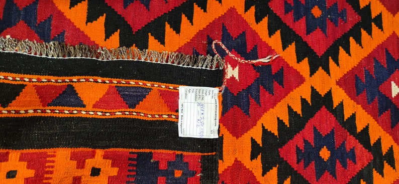 May include: A close-up of a colorful rug with a geometric pattern in shades of orange, red, black, and blue. The rug has a fringe border and a price tag attached to it.