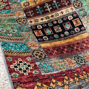 K&ouml;nnte beinhalten: Ein bunter Teppich mit einem geometrischen Muster in Rot-, Blau-, Gr&uuml;n- und Goldt&ouml;nen. Der Teppich hat einen Fransenrand und ist aus einem weichen, gewebten Material gefertigt.