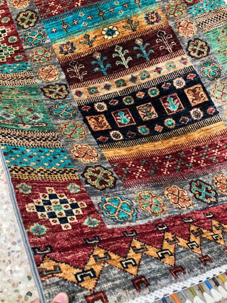 K&ouml;nnte beinhalten: Ein bunter Teppich mit einem geometrischen Muster in Rot-, Blau-, Gr&uuml;n- und Goldt&ouml;nen. Der Teppich hat einen Fransenrand und ist aus einem weichen, gewebten Material gefertigt.