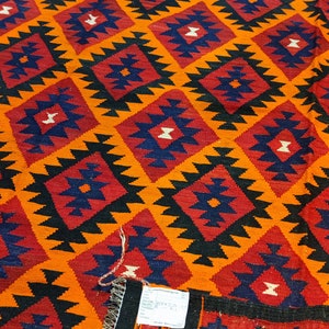 May include: A colorful rug with a geometric pattern in shades of orange, red, black, and blue. The rug has a fringe border and a tag with the text "www.iranianrugs.com".
