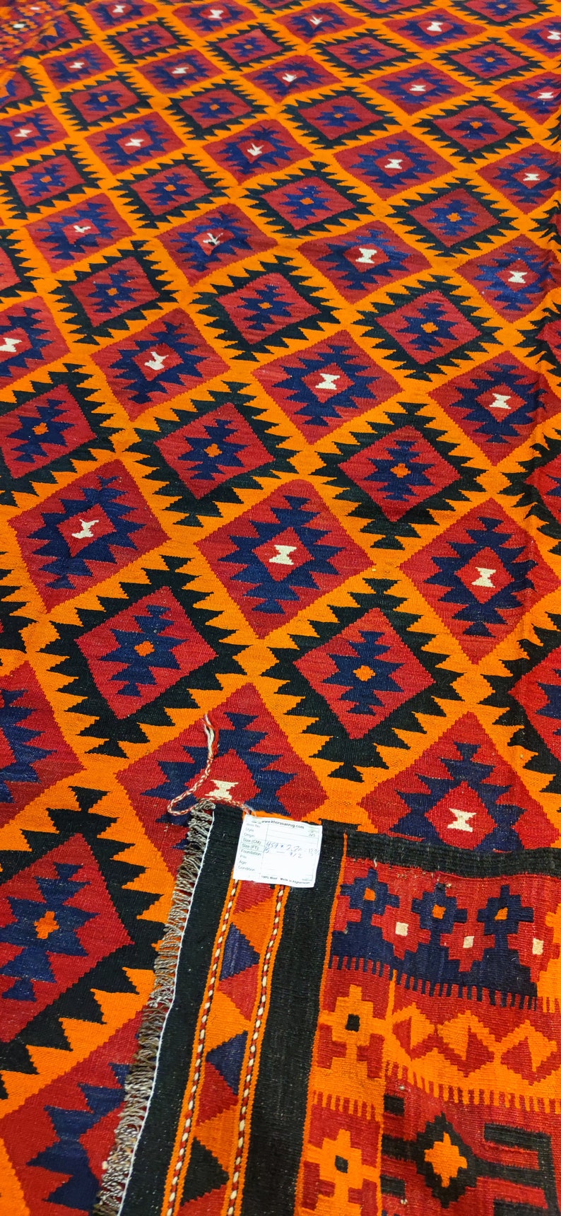 May include: A colorful rug with a geometric pattern in shades of orange, red, black, and blue. The rug has a fringe border and a tag with the text "www.iranianrugs.com".