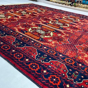 May include: A large rectangular area rug featuring a deep red field with intricate geometric patterns in navy blue, orange, and cream. The border displays a repeating floral motif in red and navy blue, with a dense lattice texture throughout.