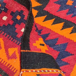 May include: Close-up of a colorful woven rug with a geometric pattern in shades of red, orange, black, and blue. The rug has a fringe border.