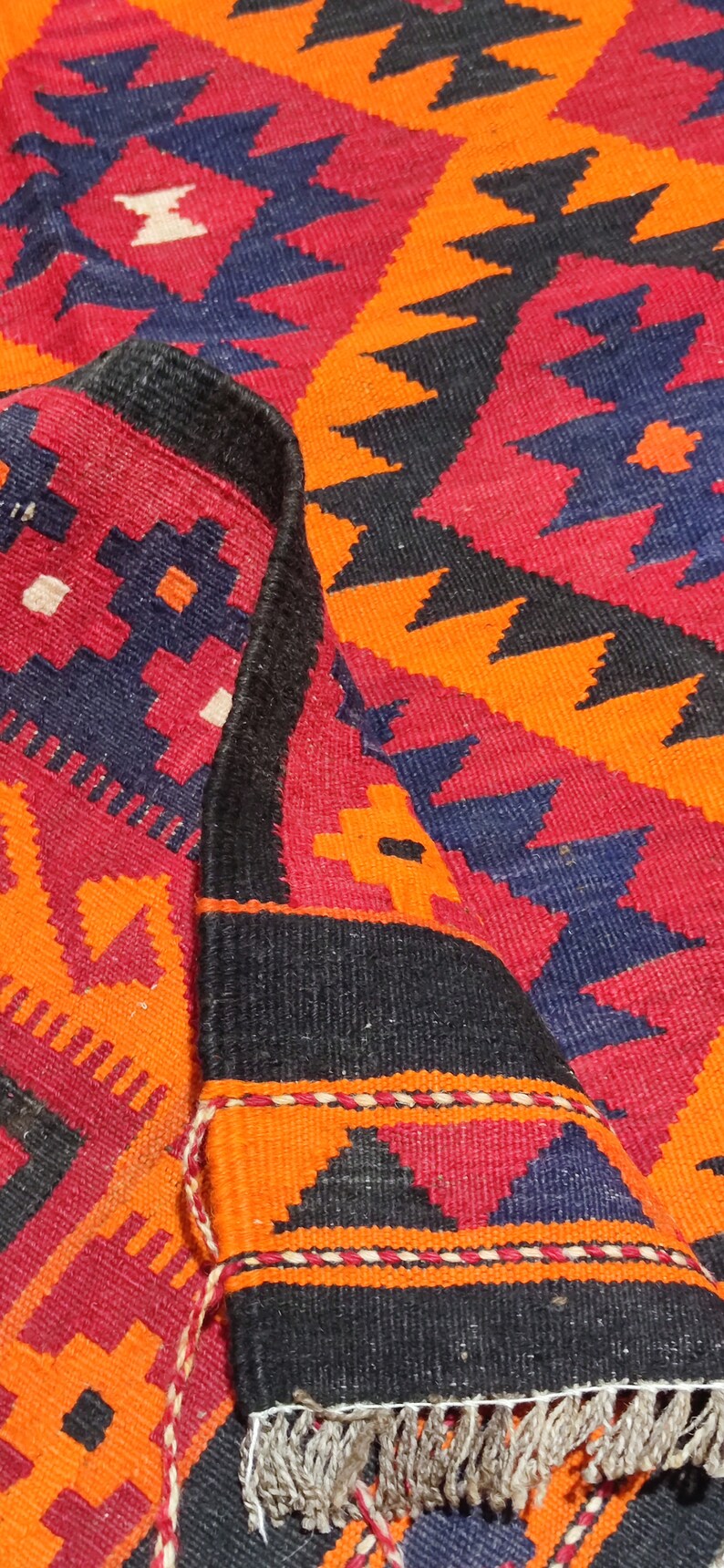May include: Close-up of a colorful woven rug with a geometric pattern in shades of red, orange, black, and blue. The rug has a fringe border.