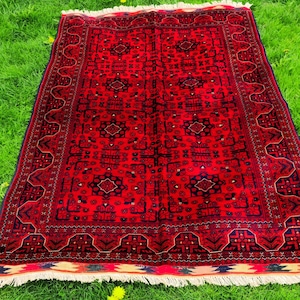 Op de afbeelding: Een rechthoekig rood tapijt met een ingewikkeld geometrisch patroon en een donkere rand. Het tapijt heeft een centraal rood veld met herhalende donkerblauwe en zwarte vormen. Het tapijt ligt op groen gras en heeft een witte franje.