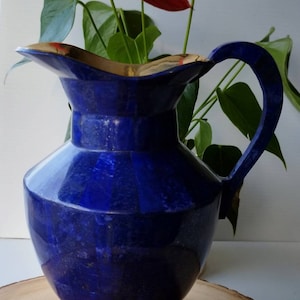 May include: A blue ceramic pitcher with a gold rim and handle. The pitcher is decorated with a textured pattern and is sitting on a wooden surface. There are red flowers in the background.