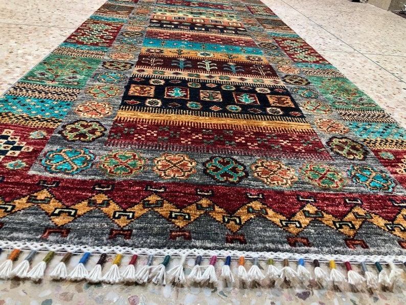 K&ouml;nnte beinhalten: Ein bunter Teppich mit geometrischem Muster und Fransen. Der Teppich hat eine Vielzahl von Farben, darunter Rot, Blau, Gr&uuml;n und Gelb. Der Teppich ist aus Wolle und hat eine weiche Textur.