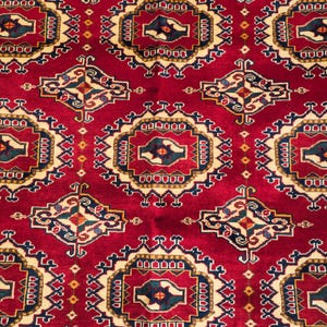 Könnte beinhalten: Nahaufnahme eines roten Orientteppichs mit einem detaillierten geometrischen Muster. Das Design zeigt sich wiederholende Medaillons in Creme, Marineblau und Gold, wodurch eine reichhaltige, strukturierte Oberfläche entsteht. Der Rand des Teppichs hat ein komplexes Blumenmuster.