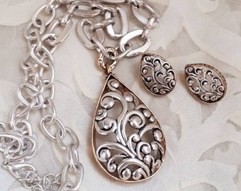 May include: A silver chain necklace with a gold and silver teardrop pendant and matching earrings. The pendant and earrings feature a delicate, intricate design of swirls and circles.