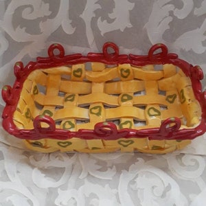 May include: A yellow ceramic bread basket with a red rim and green heart designs. The basket has a woven design and is perfect for serving bread or other snacks.