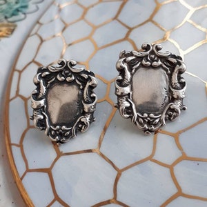 Puede incluir: Un par de pendientes de plata con un diseño ornamentado detallado. Los pendientes presentan una forma ovalada de plata rodeada por un marco decorativo.