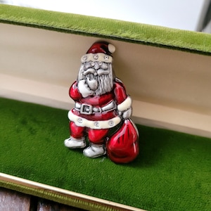 Puede incluir: Un broche de Papá Noel con traje rojo, ribetes blancos y barba plateada. El broche tiene un saco rojo y está adornado con pedrería. El pin festivo está sobre un fondo de terciopelo verde.