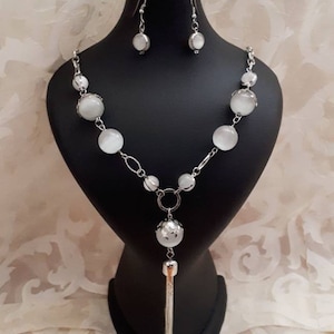 May include: A silver necklace with a white stone pendant and a tassel. The necklace is on a black display stand. There are matching earrings with white stones.