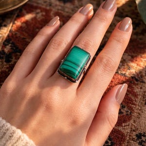 May include: A rectangular ring featuring a vibrant green malachite stone. The stone is set in a silver-toned metal frame. The ring is displayed on a hand with neutral-toned nail polish, set against a patterned textile background.