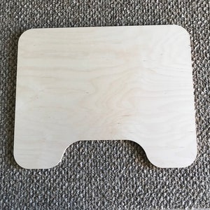 May include: A light-colored, rectangular wooden tray with rounded corners and a curved cutout on one side. The tray is made of unfinished wood and sits on a gray, textured surface.