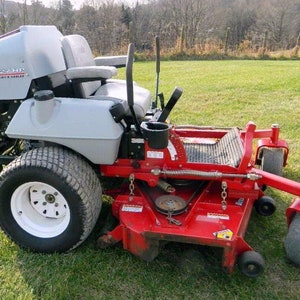 May include: A red and gray zero-turn riding lawn mower with a large grass catcher. The mower has a seat with a back rest and a cup holder. The mower is on a grassy lawn.