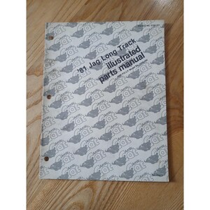 Op de afbeelding: Een open handleiding getiteld '81 Jag Long Track illustrated parts manual' met het Arctic Cat-logo herhaald over de omslag. De handleiding heeft drie gaten om te binden en ligt op een houten oppervlak.