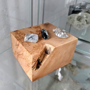 May include: A light-colored, square wooden block with a natural wood grain pattern. Three polished gemstones rest on top: a clear stone with black inclusions, a black and gray stone, and a clear, faceted stone. The block has a carved-out section on one side.