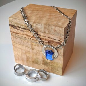 Pode incluir: Um colar prateado com um grande pingente retangular de pedra preciosa azul. O colar é exibido em um bloco de madeira, com três anéis prateados em primeiro plano. A corrente tem um design único com elos circulares e em espiral.
