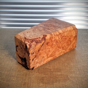 Pode incluir: Um bloco retangular de madeira com um padrão de grão natural. A madeira tem uma cor castanho-avermelhada quente com áreas mais escuras e texturizadas. As bordas do bloco são ligeiramente irregulares, mostrando a forma natural da madeira.