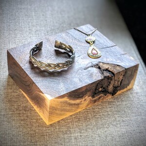 Pode incluir: Uma pulseira de punho trançada prateada e um colar com pingente com uma pedra preciosa rosa em forma de gota repousam sobre um bloco de madeira rústico. O colar tem uma corrente delicada e um pingente circular acima da gema.