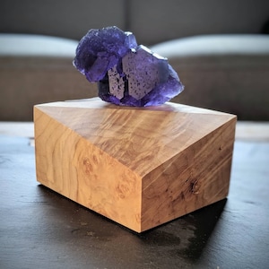 May include: A vibrant purple fluorite crystal rests atop a geometric wooden display stand. The stand is crafted from light-colored wood with a natural grain pattern. The crystal's facets catch the light, creating a sparkling effect.