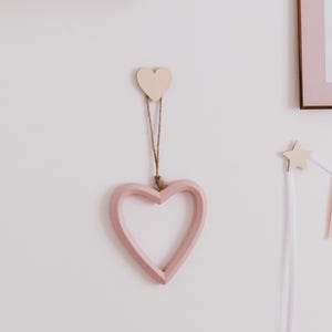 May include: A pink heart-shaped wall decoration suspended by a jute string, with a small wooden heart above. A framed pink bow print and a banner with star and triangle pennants adorn the white wall. The decor has a soft, pastel colour scheme.