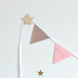 May include: A decorative banner featuring pink and rose gold glitter triangle flags strung on white ribbon. A wooden star is attached to the top, and a gold star is at the bottom. The banner is against a white background.