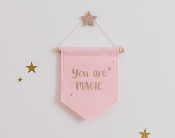 May include: A pink felt banner with the words "You are MAGIC" in gold glitter lettering. The banner is suspended from a wooden dowel and white string, with a gold glitter star above. Gold star decals are on the white wall.