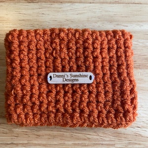 Peut inclure: Un &eacute;tui crochet&eacute; de couleur orange avec un motif textur&eacute;. Une petite &eacute;tiquette en bois avec le texte "Danni's Sunshine Designs" est attach&eacute;e. De forme rectangulaire.