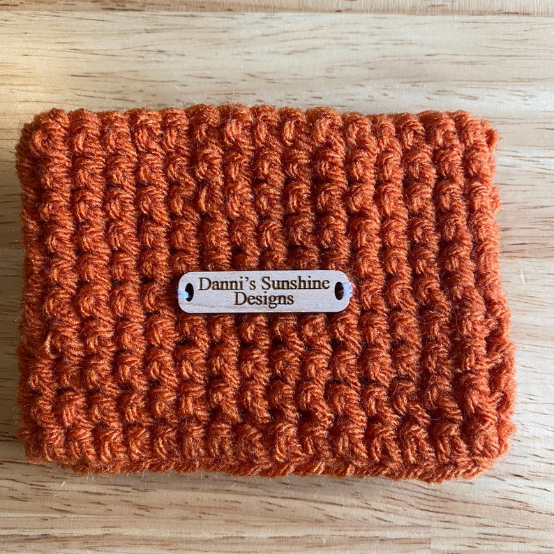 Peut inclure: Un &eacute;tui crochet&eacute; de couleur orange avec un motif textur&eacute;. Une petite &eacute;tiquette en bois avec le texte "Danni's Sunshine Designs" est attach&eacute;e. De forme rectangulaire.
