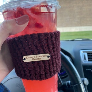 Peut inclure: Un gobelet en plastique transparent rempli d'une boisson rouge et de glace. La tasse est envelopp&eacute;e dans une housse en crochet bordeaux fonc&eacute; avec une &eacute;tiquette en bois qui dit "Danni's Sunshine Designs". La boisson est dans une voiture.