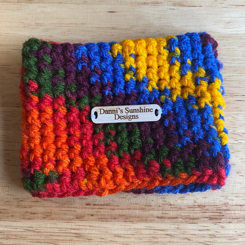 Peut inclure: Un dessous de verre crochet&eacute; &agrave; la main, aux couleurs vives, avec une &eacute;tiquette en bois portant l'inscription "Danni's Sunshine Designs". Il pr&eacute;sente un m&eacute;lange de couleurs vives, notamment orange, rouge, vert, violet, bleu et jaune.