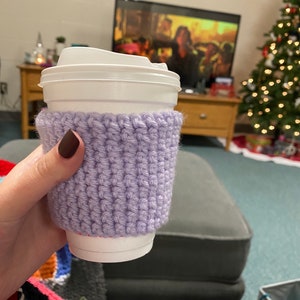 Peut inclure: Un manchon de tasse &agrave; caf&eacute; en crochet lilas. Le manchon est enroul&eacute; autour d'une tasse &agrave; caf&eacute; jetable blanche avec un couvercle blanc. Le manchon est fait d'un mat&eacute;riau tricot&eacute; textur&eacute;. L'arri&egrave;re-plan comprend un sapin de No&euml;l et une t&eacute;l&eacute;vision.