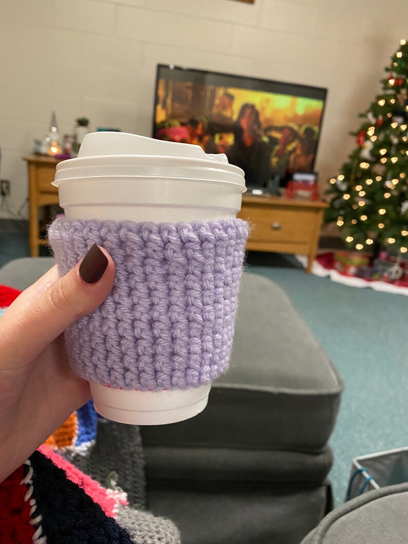 Peut inclure: Un manchon de tasse &agrave; caf&eacute; en crochet lilas. Le manchon est enroul&eacute; autour d'une tasse &agrave; caf&eacute; jetable blanche avec un couvercle blanc. Le manchon est fait d'un mat&eacute;riau tricot&eacute; textur&eacute;. L'arri&egrave;re-plan comprend un sapin de No&euml;l et une t&eacute;l&eacute;vision.